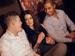 Two smiling men in white shirts chat with a dark-haired woman between them on a lounge sofa at a party