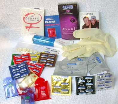 Assorted condom packets, latex gloves, dental dams, and lube sachets spread out on a white surface