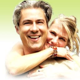Smiling blonde woman hugs a grey-haired man from behind, both cheerful against a soft green background