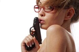 Blonde woman in red glasses holds a black handgun close to her face, shoulder bare, white background
