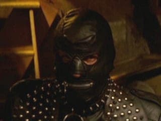 Person wearing a black leather hood and studded harness sits in dim light, a BDSM-styled portrait