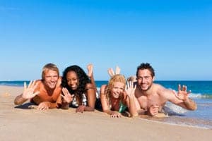 Two couples in swimwear lying on a sunny beach at the water's edge, smiling and waving at the camera