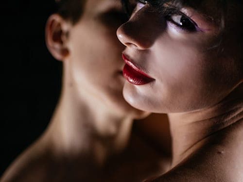 Close-up studio portrait of a woman with dark hair and red lipstick beside a partner, dramatic low lighting