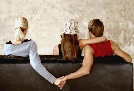 Rear view of three people on a leather couch, a man hugs a woman in red while secretly holding another woman's hand