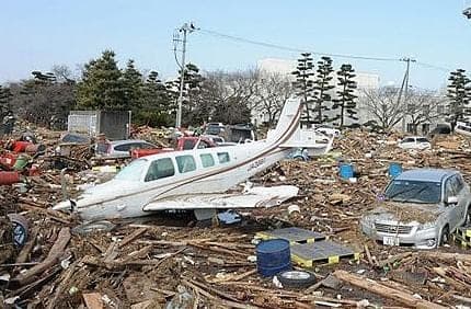 Tsunami aftermath with a small white airplane marooned among wrecked cars and scattered debris on muddy ground