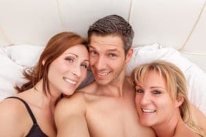 Smiling man lying between two women on white bed linens, all three looking up at the camera in a close portrait