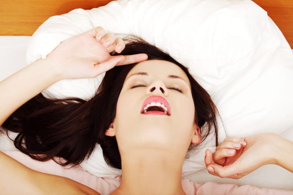 Dark-haired woman lying on white bedding with head tilted back, eyes closed and mouth open in pleasure