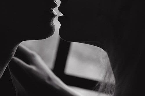 Black and white close-up of two people about to kiss, lips nearly touching in soft window light