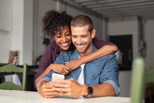 Smiling couple at a kitchen table, woman hugging man from behind as they look at a smartphone together