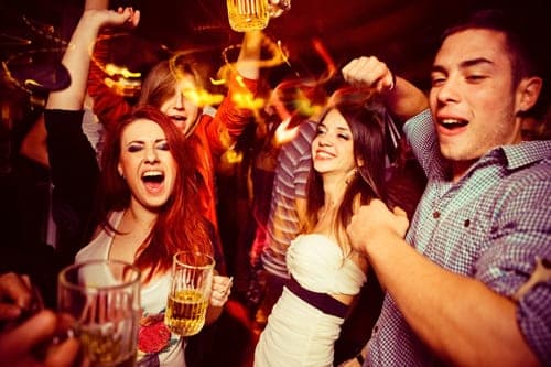 Young people laughing and dancing in a crowded nightclub, holding beer mugs under warm stage lights