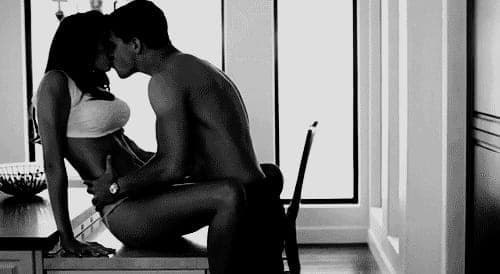 Black and white photo of a shirtless couple kissing while seated on a kitchen counter by a bright window