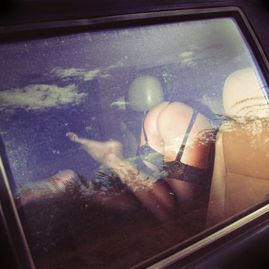 View through a car window of a woman in black lingerie and fishnets reclining across the back seat