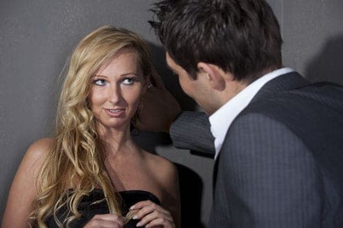 Blonde woman in a black dress looking up at a man in a grey suit against a dark wall