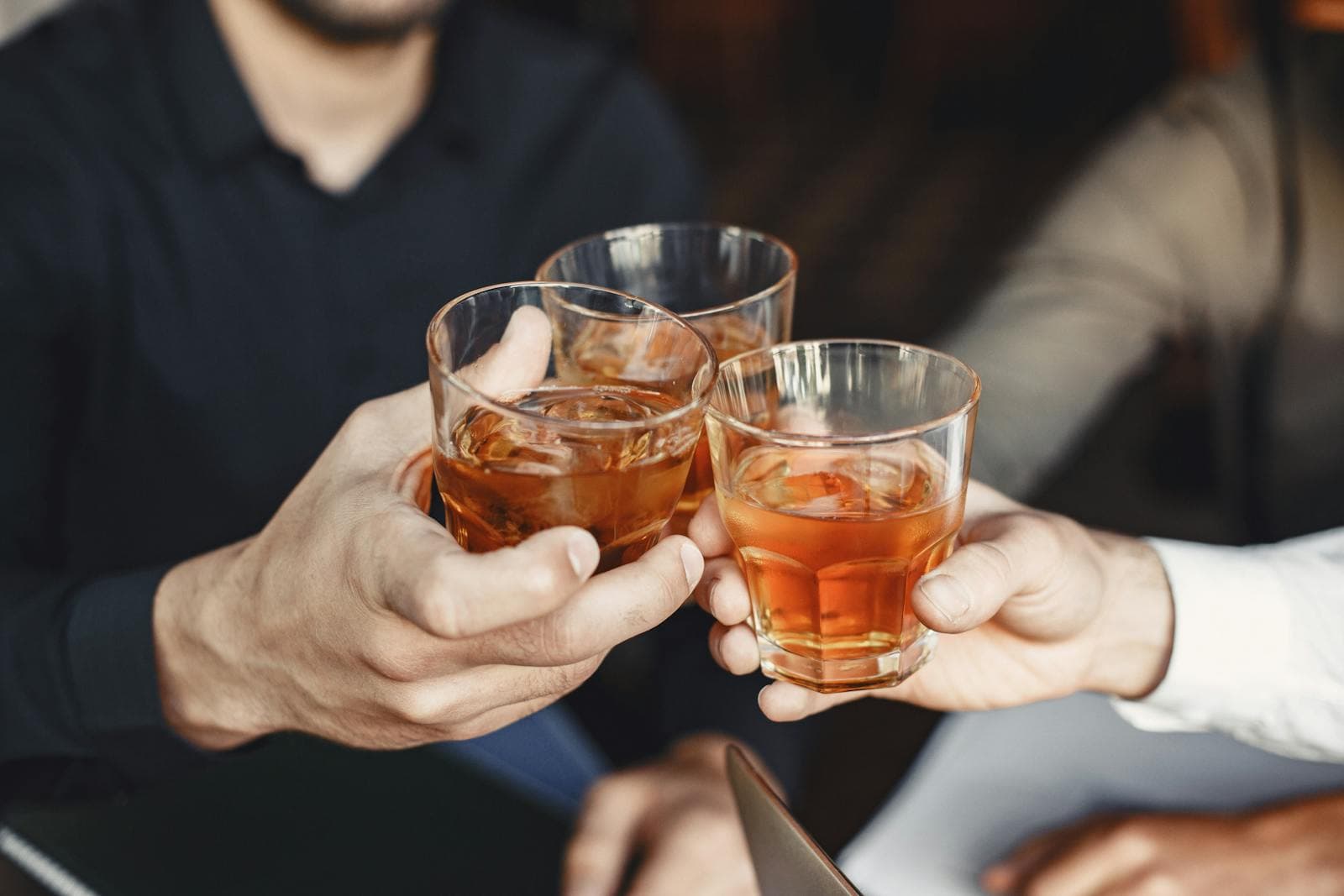 Three people clinking whiskey glasses together in a celebratory toast at an indoor wooden table