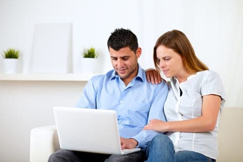 Young couple sitting on a white sofa smiling while looking at a laptop together