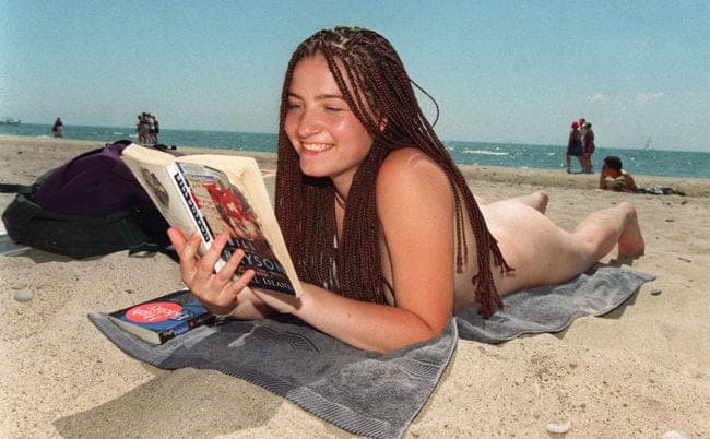 Smiling woman with long braided hair reading a book while sunbathing on a sandy beach