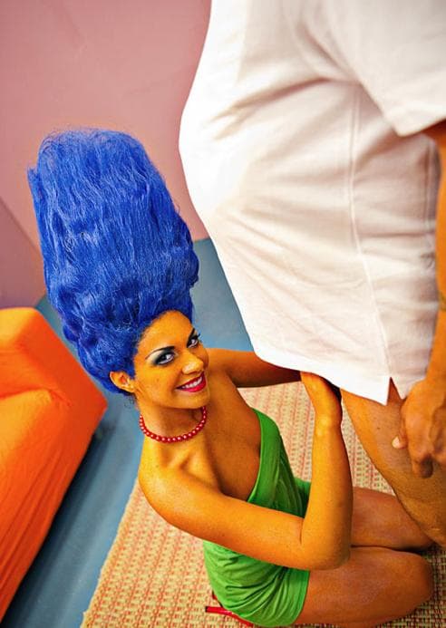 Woman in a tall blue beehive wig and green dress kneeling in a costumed parody scene