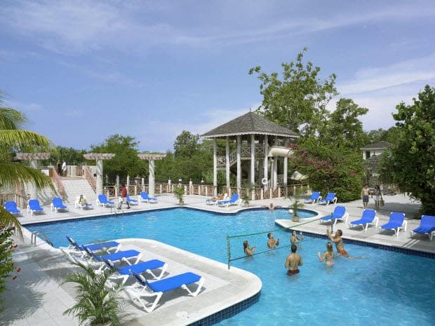Tropical resort pool with blue loungers, a gazebo, and guests playing volleyball in the water