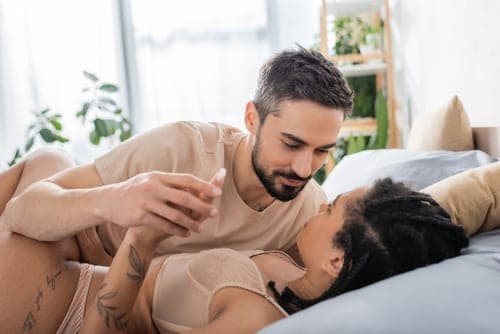 Bearded man and a woman lie face to face on a bed, smiling softly in a bright bedroom with plants
