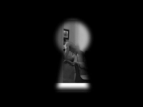 Black-and-white keyhole view into a dim room showing a blurred silhouette of a figure on a chair