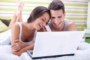 Smiling couple in white tank tops lie on a bed together looking at an open laptop screen