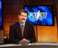 Mustachioed news anchor in a suit sits at a studio desk in front of a 20/20 program screen