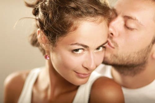 Close-up of a woman with an updo smirking at the camera as a man kisses her temple from behind